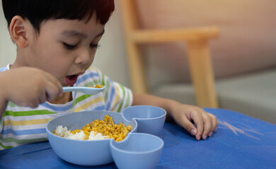 Cute Asian child eating breakfast and using spoon at home. Kid eating healthy food in kindergarten...