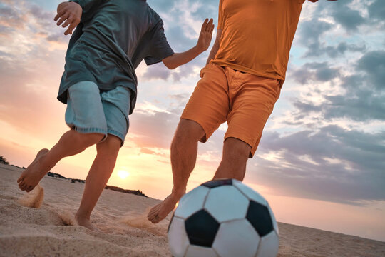 Father And Son Playing Football, Family Fun Outdoors Players In Soccer In Dynamic Action Have Fun Playing Football In The Beach, Summer Day Under Sunlight.
