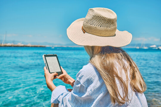 Fashion pretty woman outdoors lifestyle watching, reading on tablet ebook on the beach in summer day. Wearing wide brimmed hat, Sunbating with uv protection. Concept of beach vacation.

