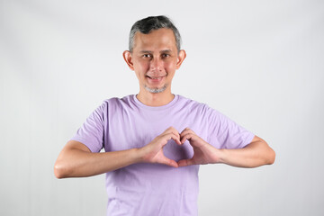 Portrait of Asian man wearing purple tshirt smiling with love sign (heart with fingers) on his chest, health concept.
