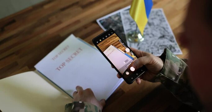 Military officer photographs top secret documents about Ukrainian counteroffensive on phone. Soldier works with papers sitting at table with yellow and blue flag
