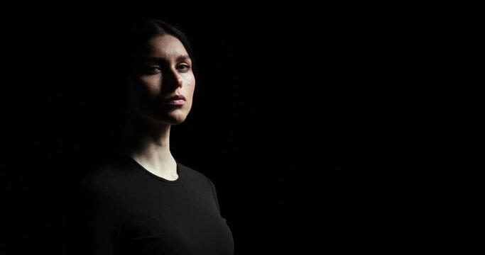 Woman of dramatic appearance stands in the dark on a black background, slowly raising her head and staring into the camera with a serious expression on her face. The shot is slow and deliberate.