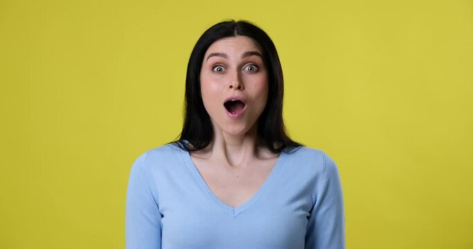 A caucasian woman stands on a yellow background, looking at the camera. Suddenly, she sees something amazing and exclaims Wow with a big smile on her face, followed by laughter.