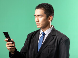 A portrait of an Asian man wearing a white shirt and a black suit, looking very happy while looking at a cellphone, isolated on a green background.