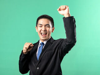 A portrait of an Asian man wearing a white shirt and a black suit, seen celebrating and raising both hands, isolated on a green background.