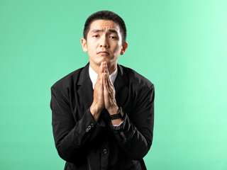 A portrait of an Asian man wearing a white shirt and a black suit, posing in apology, isolated on a green background.