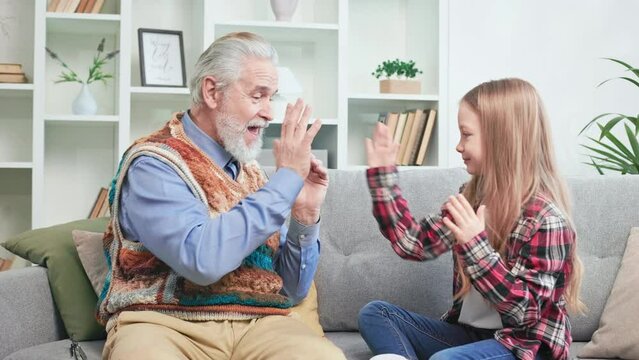 Kind granddad and joyful granddaughter enjoying each other's company on cozy couch. Retired man and girl in casual clothes playing hand-clapping game together. Concept of two generations and love.