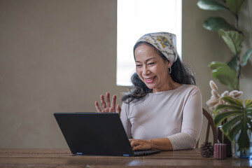 Attractive beautiful mature Asian woman sits at a table with her use laptop in her minimal living room. Hobby and lifestyle concepts