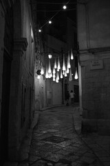 Grayscale vertical shot of a narrow stone ally at night with hanging lights
