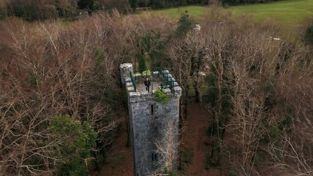 Circling drone shot of two people on top of an ancient castle tower surrounded by trees.