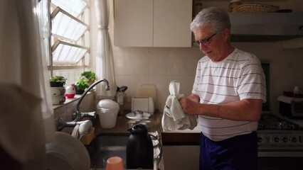 Retired senior man drying kitchenware standing at kitchen sink. Domestic lifestyle scene of older person doing home routines - Powered by Adobe