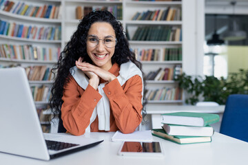 Young hispanic woman studying in academic university library, female student smiling and looking at camera while sitting at laptop, woman with curly hair. - obrazy, fototapety, plakaty