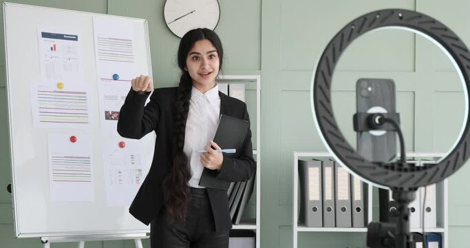 Indian businesswoman confidently stands next to a whiteboard, conducts a video call. She shares insights and points to the board. She gives a thumbs-up, and bids farewell to the participants.