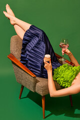Woman lying on armchair, drinking rose wine and eating sweet muffin again green background. Relaxation. Concept of pop art photography, creative vision, imagination, party. Minimal art