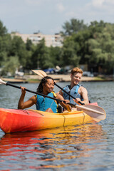 carefree interracial couple in life vests holding paddles and looking at clear water while sailing in sportive kayak during recreation weekend on summer day