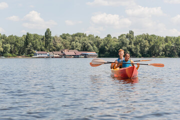 Fototapeta na wymiar cheerful african american woman and young redhead man in life vests paddling in sportive kayak while spending time on river during weekend on summer day