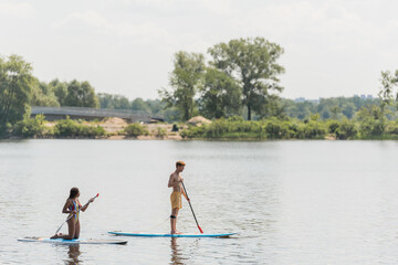 Fototapeta na wymiar side view of young sportive man and african american woman standing on knees and sailing on sup boards with paddles on river with green bank in summer