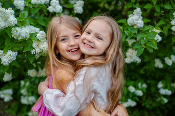 two happy blonde girls with long hair hug and laugh children's friendship park lilac bush