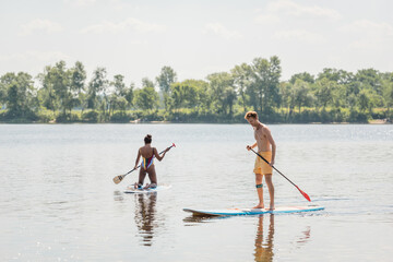 young and sportive redhead man standing on sup board with paddle near african american woman in colorful swimsuit sailing on picturesque lake with green shore