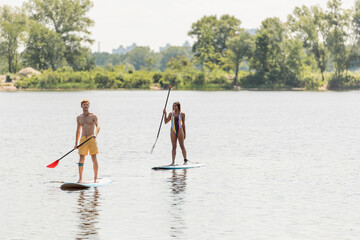 redhead man in swim shorts and african american woman in colorful swimsuit holding paddles while sailing on sup boards along green shore on summer day