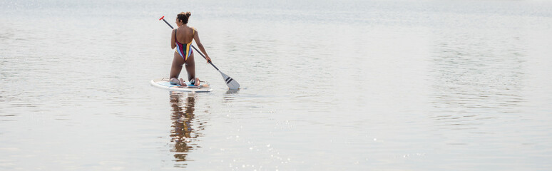 back view of african american woman in colorful and striped swimsuit spending time on lake by sailing on sup board with paddle, banner, outdoor activity, summer vibes