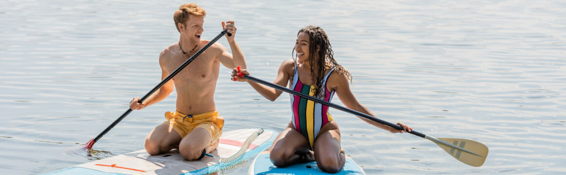 young and happy redhead man and excited african american woman in striped swimsuit kneeling on sup boards while sailing on lake during water recreation in summer, banner