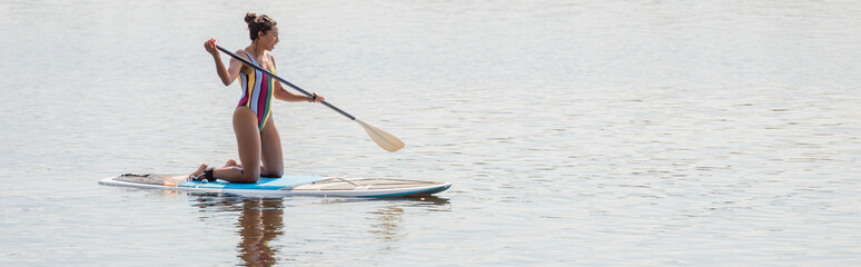 full length of sportive african american woman in colorful swimsuit holding paddle and standing on knees while sailing on sup board on lake in summer, banner