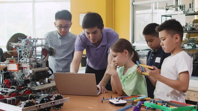 Young teacher explains to children in a class at a robotics lesson how to program a mechanical module on a laptop. Concept of technological modern education