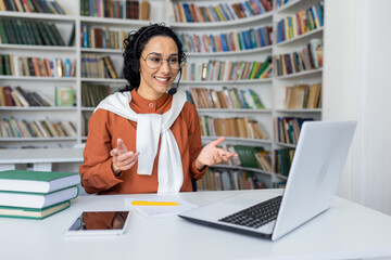 Teacher conducts lessons online, woman with video call headset smiling and explaining lecture online using laptop for remote communication and learning, sitting at desk in university library at desk.