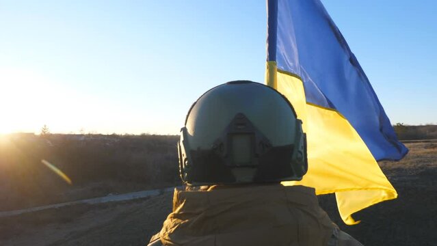 Ukrainian army girl holds lifting national banner against background of sunset. Young female soldier in military uniform stands with waving flag of Ukraine to peak of hill. Victory of war. Dolly shot
