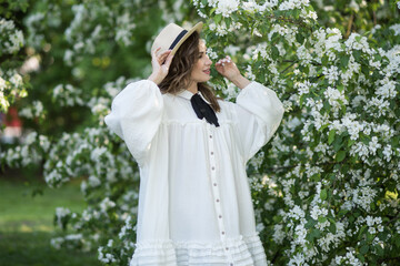 Portrait of a beautiful young girl in a hat. A girl poses near a blooming white tree. She enjoys the smell of flowers in the apple orchard.