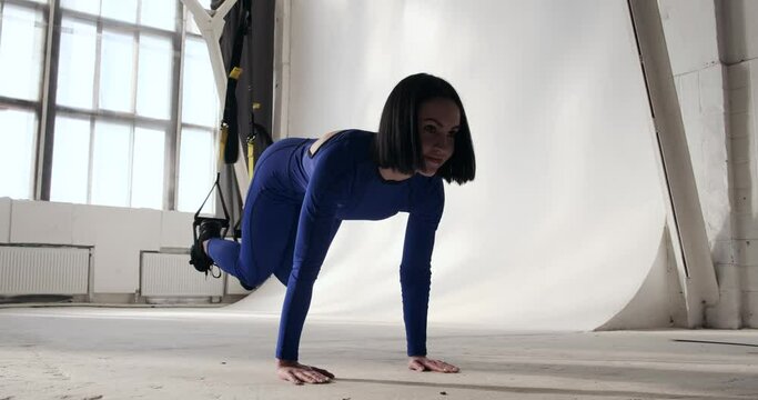 Confident Caucasian woman is wearing a blue sports jumpsuit and athletic shoes. She is performing core-strengthening exercises in a plank position using fitness bands with loops attached to her legs.