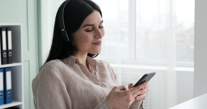 Cheerful businesswoman wearing wireless headphones is typing on her phone and chatting with friends while listening to music. She is having fun and enjoying her break time.