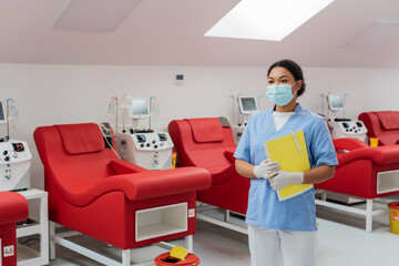 multiracial healthcare worker in uniform, medical mask and latex gloves standing with paper folder...