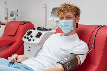 Fototapeta na wymiar redhead donor in medical mask and blood pressure cuff looking at camera while sitting on ergonomic chair near automated transfusion machine in hospital