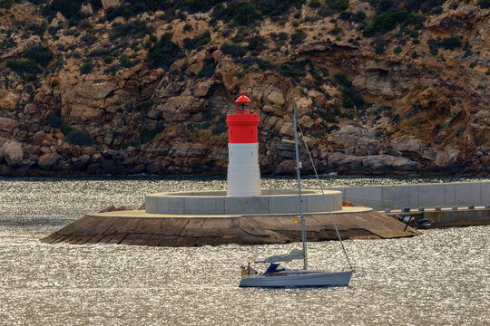 A sailing yacht enters the port of Cartagena. In the background is a red lighthouse. The waves glitter in the sunlight.