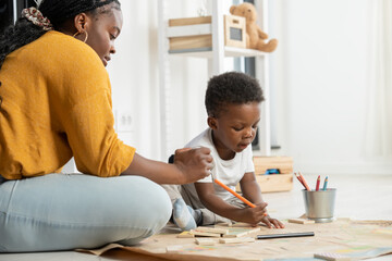 Cheerful African-American mother and cute toddler son playing with colored pencils, drawing on a big piece of paper, and sitting on the floor. Mother-son fun bonding time. Daycare creative activities. - obrazy, fototapety, plakaty