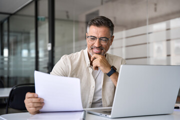 Smiling Latin or Indian male business man accountant analyst holding documents, work at laptop computer doing online trade market tech research. Focused Hispanic businessman with paperwork in office. - obrazy, fototapety, plakaty