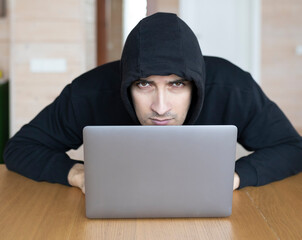 Portrait of enigmatic man in hoodie sits at the desk and works on laptop	