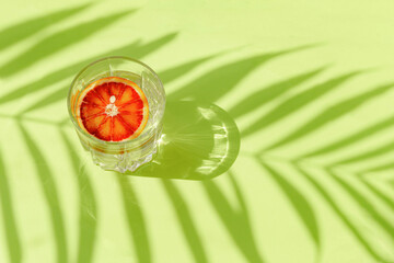Glass of water with sliced blood orange on pastel green background with palm tree leaf shadow. Summer refreshment and party concept. Minimal style.