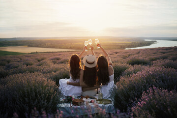Girlfriends having picnic in the lavender field at sunset. Group of young women sitting on lavender field on summer day. Girlfriends drinking wine on outdoor party. - obrazy, fototapety, plakaty