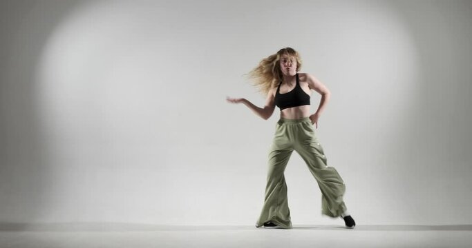Curly-haired Caucasian dancer on white background as she unleashes her jazz funk skills, with dynamic movements and infectious energy. She wearing green pants and black top.