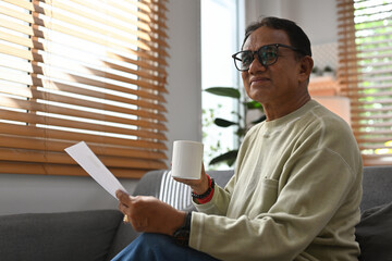 Portrait with middle aged man looking forward and holding a cup of coffee and paper document while sitting on the sofa.