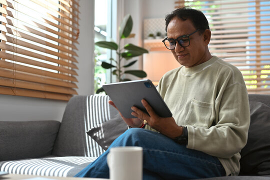 The image features middle aged Asian man wearing a long-sleeved sweater sitting on a sofa looking relaxed and using digital tablet at home.