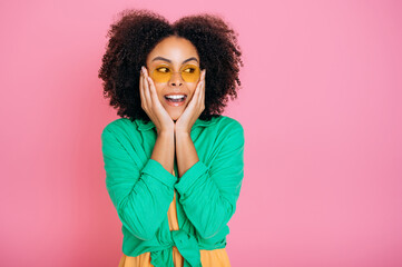 Excited positive african american or brazilian curly haired pretty trendy woman in green shirt, amazed looking aside, holding palms near cheeks and opening her mouth, stand on isolated pink background