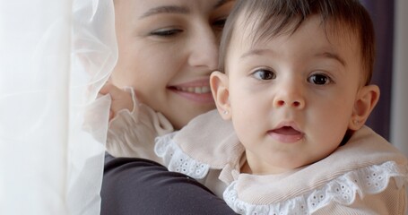 The mental balance mother baby. Portrait of a baby dark hair and mother natural light. She waves her hand in greeting. A smile on a woman's face. Earring in the ear.