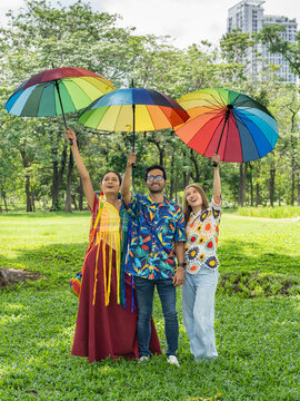 young diverse friends raising rainbow umbrellas in hand standing laughing together, LGBTQ people enjoy weekend outdoor activities at city public park during summer day in pride month