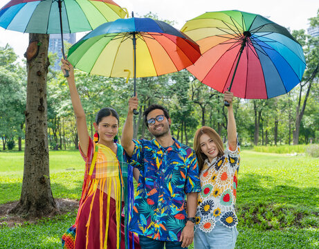 young diverse friends raising rainbow umbrellas in hand standing looking at camera, LGBTQ+ people enjoy weekend outdoor activities at city public park during summer day in pride month