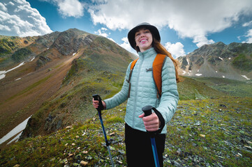 Portrait, the girl is happy and smiling while hiking in the mountains. Beautiful young caucasian sporty woman joyful stands with a backpack and looks at the camera - obrazy, fototapety, plakaty