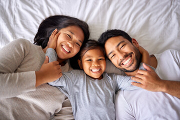 Portrait, love and family in bed from above happy, smile and bonding in their home together. Face, girl child and top view of parents with their daughter in a bedroom, relax and embracing indoors